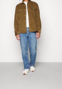 Chaqueta de camisa de pana marrón con dos bolsillos en el pecho, combinada con jeans rectos de color azul claro y zapatillas blancas, de pie contra un fondo neutro.
