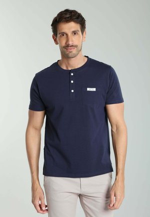 Homme portant un t-shirt henley bleu marine à manches courtes avec trois boutons et un pantalon beige, debout sur un fond gris clair.