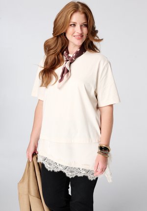 Femme aux longs cheveux roux portant un haut crème avec un ourlet en dentelle, un pantalon noir, un foulard à pois autour du cou, des bracelets, et tenant une veste beige.