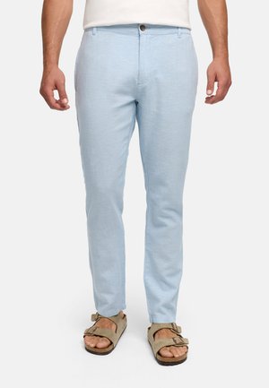 Mężczyzna noszący jasnoniebieskie wąskie spodnie i beżowe sandały na dwa paski, stojący na jednolitym białym tle.