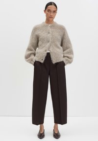 Femme avec les cheveux en chignon portant un cardigan beige en peluche à boutons, un pantalon large noir et des chaussures pointues noires, se tenant avec les mains dans les poches.