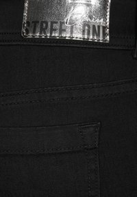 Vaqueros de mezclilla negra con una etiqueta plateada marcada "Street One". La tela es suave, con un diseño limpio y simple, y detalles de costura mínimos.
