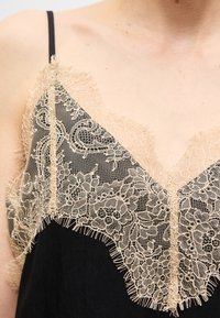 Μαύρο ένδυμα με μπεζ φλοράλ δαντέλα κατά μήκος της γραμμής του λαιμού και λεπτό μαύρο λουράκι ώμου, που δείχνει το επάνω μέρος του στήθους και της περιοχής των ώμων.