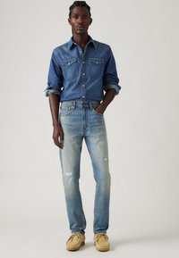Cămășă din denim cu două buzunare pe piept, blugi albaștri cu detalii uzate și pantofi mocasini bej. Modelul stă în fața unui fundal deschis.