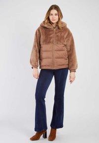 Brązowa kurtka z imitacji futra z kapturem, zapinana na zamek błyskawiczny, z puchatą dolną częścią, zestawiona z granatowymi, prążkowanymi, rozszerzanymi spodniami oraz brązowymi sznurowanymi butami na obcasie.