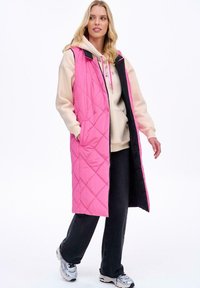 Długa pikowana kamizelka w jasnoróżowym kolorze z czarnym wnętrzem, z kieszeniami, kapturem i wzorem w diamenty, noszona na beżowej bluzie z kapturem i ciemnych spodniach.