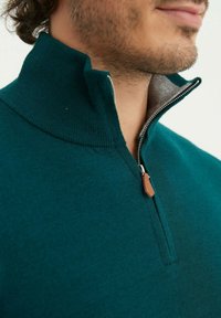 Pull-over teal avec un zip quart, texture douce, col côtelé et un tirant en cuir, mettant en valeur une doublure intérieure contrastante.