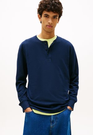 Nooruk kõverate juustega, seljas tumesinine pikade varrukatega henley-särk helerohelise t-särgi peal ja sinised teksad, käed taskutes, neutraalne taust.