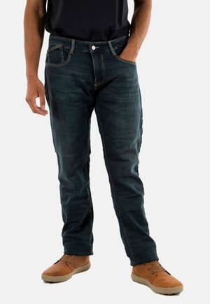 Jeans de denim azul oscuro con un diseño ajustado, cinco bolsillos y pasadores para cinturón; combinados con zapatos casuales de color marrón claro.