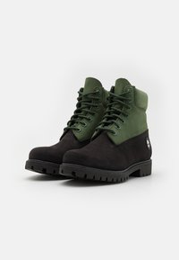 Mustad mustad ja rohelised pahkluusaapad, mis on valmistatud sujuvatest ja tekstureeritud materjalidest, paeltega sulgemisega, kummist tallaga ja bränditud detailiga küljel.