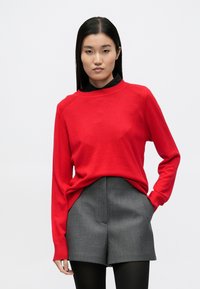 Κόκκινο πλεκτό πουλόβερ με στρογγυλή λαιμόκοψη, σε συνδυασμό με γκρι σορτς. Διαθέτει μακριά μανίκια και ριμπ μανσέτες. Φορεμένο πάνω από μαύρο πουκάμισο.