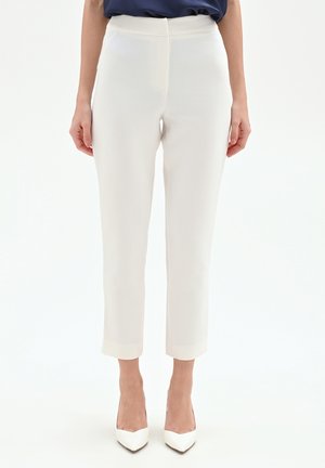 Donna che indossa pantaloni a gamba dritta a vita alta color avorio chiaro abbinati a décolleté a punta dello stesso colore su sfondo bianco.