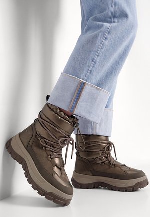 Personne portant un jean bleu clair à revers et des bottes d'hiver marron métallisé à lacets avec des semelles épaisses crantées sur fond blanc.