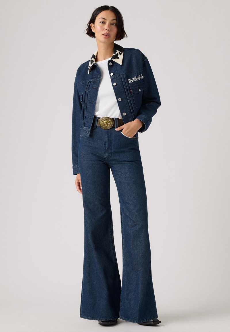 Denimjakke med krave i kvægprint, mørkeblå flared jeans og et bredt bælte med et prægede cirkulært spænde, sat mod en neutral baggrund.