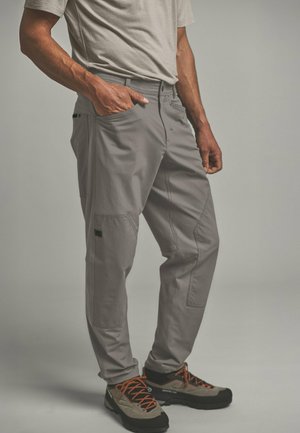 Mann trägt hellgraue Cargohosen und beige Wanderschuhe mit orangen Schnürsenkeln, steht mit einer Hand in der Tasche vor einem einfarbigen Hintergrund.