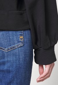 Pullover nero con maniche a sbuffo accanto a jeans in denim blu con cuciture dorate e un piccolo logo ricamato sopra la tasca.