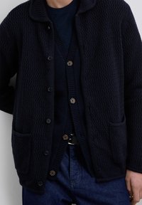 Homme portant un cardigan en tricot navy foncé à superposition et une veste texturée sur un jean bleu avec une ceinture noire, se tenant contre un fond clair.
