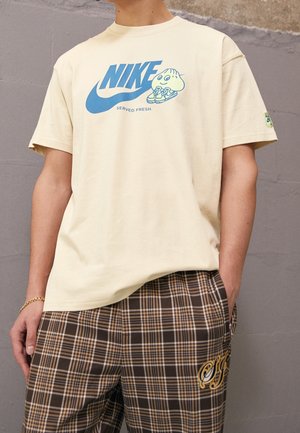 Beżowa koszulka Nike z niebieskim logo, kreskówkowy pieróg trzymający buty, napis "Served Fresh", noszona z brązowymi kraciastymi spodniami z haftowanymi detalami.