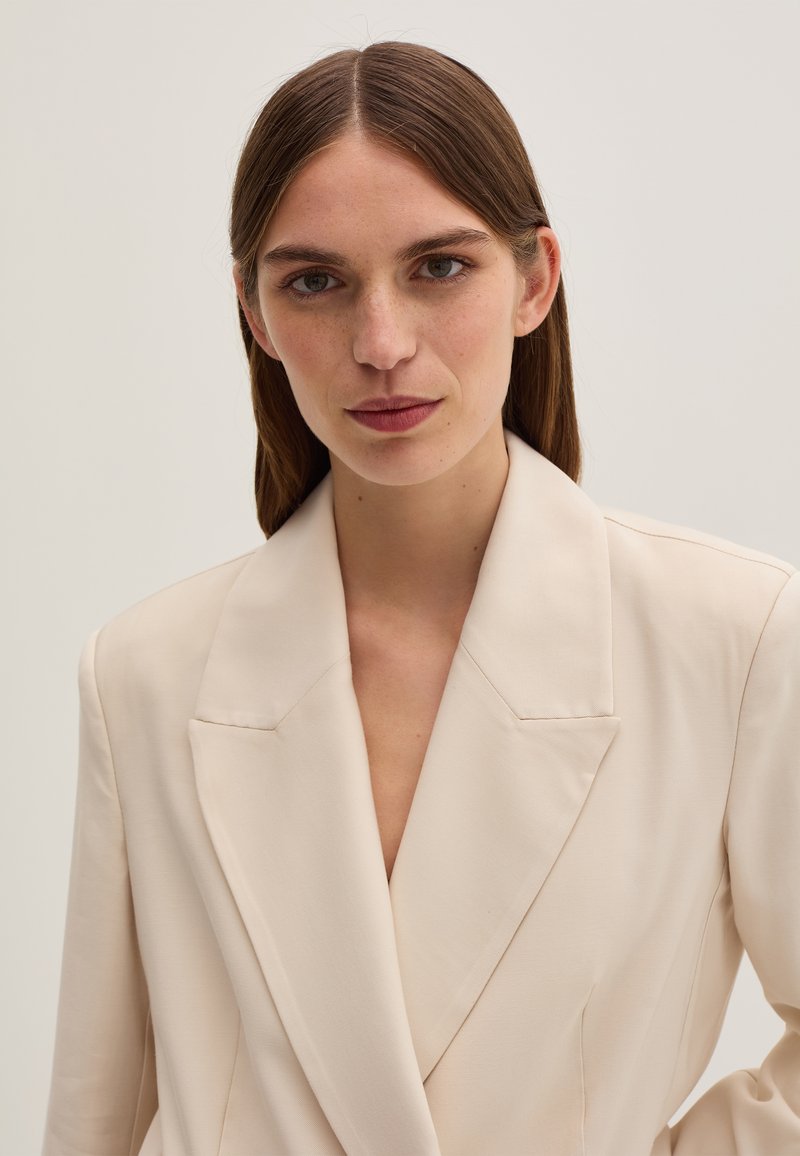 Blazer color crema con scollatura profonda a V, spalle strutturate e ampi revers. Il tessuto è liscio con una finitura leggermente testurizzata.
