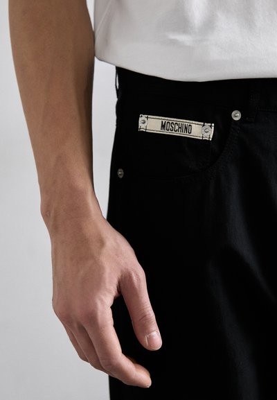 Svarta bomullsjeans har en vit Moschino-logga på fickan, silverdetaljer och en slim fit-design, visade med en hand som håller i midjan.