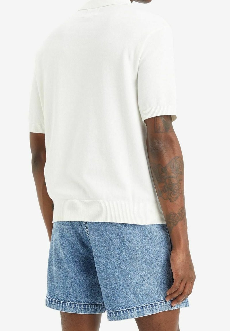 Homme portant un t-shirt blanc uni à manches courtes et un short en jean bleu clair, vu de dos avec des tatouages visibles sur les bras.