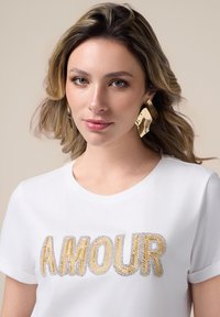 Camiseta corta de manga blanca con bordado de "AMOUR" en hilos dorados y plateados. La modelo lleva un pendiente texturizado dorado.