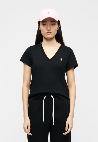 Νεαρή γυναίκα που φοράει μαύρο T-shirt με V-λαιμόκοψη και μαύρο παντελόνι με λευκά κορδόνια, και ένα ανοιχτό ροζ καπέλο μπέιζμπολ με λογότυπο.