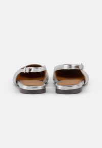Sandalias metálicas plateadas con la parte trasera abierta, tirante en el tobillo y una suela interior marrón. Presentan una suela plana y un diseño minimalista.