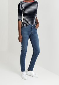 Navy and white striped long-sleeve top with a red neckline, paired with blue slim-fit jeans and white sneakers, standing on a light background.