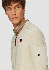 Beige gestrickte Zip-Jacke mit einem strukturierten Oberteil, gerippten Akzenten und einem Lederlogo-Patch. Über einem braunen karierten Hemd getragen.