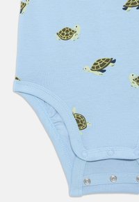 Valkoinen bodysuit, jossa on useita kilpikonnakuoseja vihreinä ja ruskeina. Siinä on nepparit alaosassa ja pehmeä kangas.