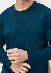 Miętowo-zielony, dziergany sweter z okrągłym dekoltem, ściągaczami na rękawach i gładką fakturą. Na pierwszym planie tors i dłonie.