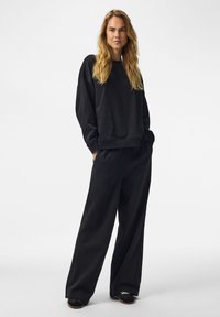 Femme aux longs cheveux blonds vêtue d’un sweat-shirt noir ample et d’un pantalon noir à jambes larges, debout les mains dans les poches sur un fond blanc.