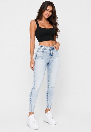 Frau trägt ein schwarzes Croptop, hellblaue, hoch taillierte Skinny-Jeans und weiße Sneakers und steht vor einem einfarbigen Hintergrund.