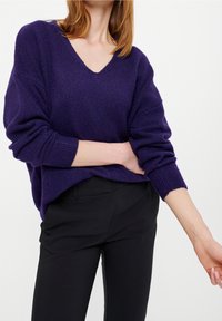 Femme portant un pull ample violet foncé à col en V avec manches longues et un pantalon noir ajusté, sur un fond clair uni.