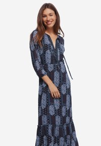 Uśmiechnięta kobieta z długimi brązowymi włosami, ubrana w długą sukienkę w granatowo-paisleyowy wzór z dekoltem w kształcie litery V i rękawami do trzech czwartych.