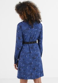 Blaues Kleid mit einem gepunkteten Muster in Schwarz und Weiß, langen Ärmeln und einem taillierten schwarzen Gürtel in der Taille. Strukturierter Stoff.