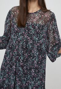 Šaty s květinovým vzorem a černým průhledným překryvem. Mají krátké rukávy, kulatý výstřih a volný střih s barevnými detaily v růžové a zelené barvě.