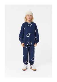 Completo felpa e pantaloni blu navy con design di faccine sorridenti bianche, realizzato in tessuto morbido. Abbinato a un berretto lavorato a maglia beige e sneakers bianche.
