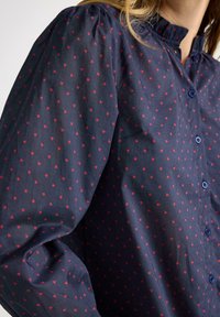 Námořnicky modrá košile na knoflíky s červeným vzorem srdcí, která má řasený design na ramenech a stojáček, vyrobená z hladké látky.