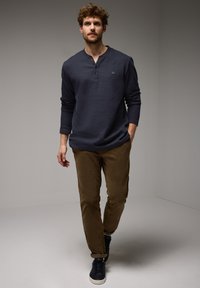 Námořnická modrá košile s dlouhým rukávem typu henley s knoflíkovou légou, kombinovaná s hnědými kalhotami a tmavými teniskami. Jednoduchý design, hladká textilie.
