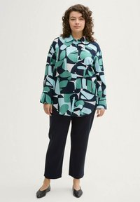Chemise à boutons avec un fond bleu marine ornée de grandes formes géométriques vertes et bleues ; associée à un pantalon foncé et des ballerines noires.