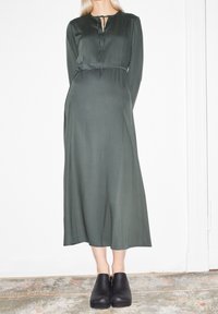 Tummanvihreä, pitkähihoinen mekko pyöreällä pääntiellä, etuosan aukolla ja vyötäröllä solmittavalla yksityiskohdalla, yhdistettynä mustiin puukenkäihin. Sileä pinta.