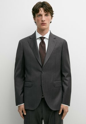 Giovane uomo che indossa un abito a righe grigio scuro, camicia bianca e cravatta marrone scuro, in piedi contro uno sfondo bianco semplice.
