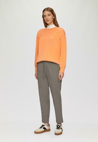 Oranje sweatshirt met het reliëfwoord "JOYFUL", grijze cargobroek met zijzakken en beige sneakers met zwarte en bruine accenten.