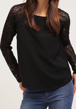 Schwarze Bluse mit durchsichtigen Spitzen-Langärmeln, Rundhalsausschnitt und glattem Stoffkörper. Kombiniert mit blauen Jeans, ohne Muster oder Akzente am Körper.