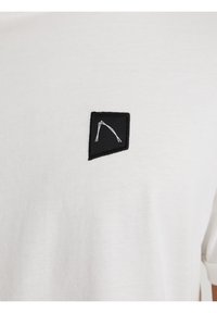 Weißes Baumwoll-T-Shirt mit einem schwarzen Stoffpatch, das mit einem gestickten geometrischen Design verziert ist. Rundhalsausschnitt und kurze Ärmel.