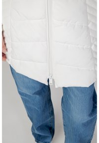 Doudoune blanche avec des coutures horizontales et une fermeture éclair sur le devant, associée à un jean en denim bleu. Le tissu semble lisse et léger.