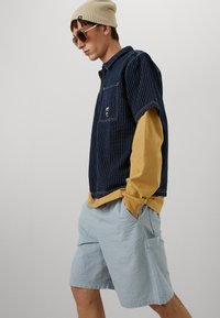 Camicia a maniche corte blu con righe, indossata sopra una maglietta lunga gialla, accompagnata da pantaloni corti azzurri con texture e completata con occhiali da sole e un berretto.
