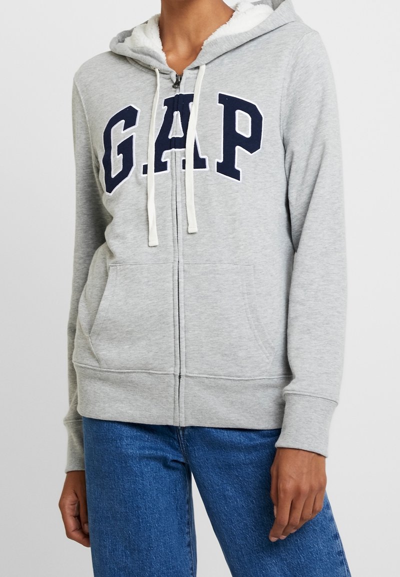 Persona vistiendo una sudadera con cremallera gris que tiene "GAP" en letras azul marino, con cordones blancos y vaqueros azules, de pie contra un fondo uniforme.